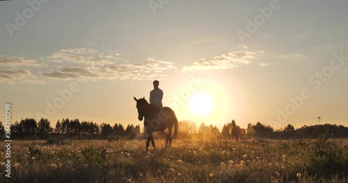 Steadicam movement of the camera around a rider in a white shirt galloping on a brown horse across the morning field lit by the dawn sun, steam is coming from the horse's nostrils, two bareback horses