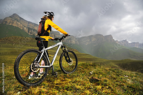 a girl in a helmet and with a backpack with a bicycle stands against the backdrop of epic mountains and rocks and cloudy evening sky. Cyclist looks at mountains before mtb descent