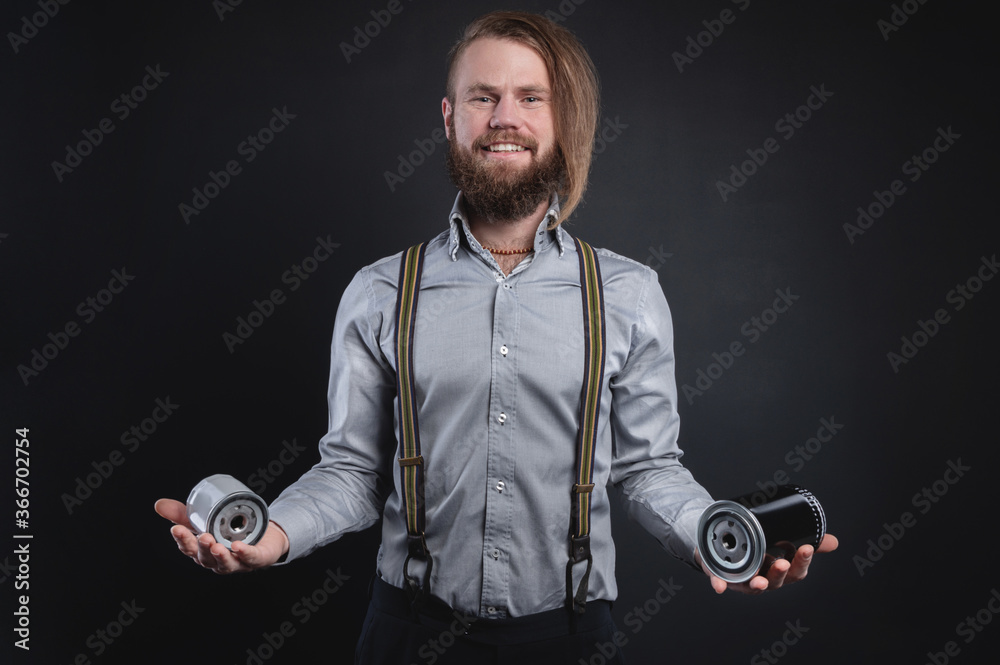 Fototapeta premium Portrait of a stylish bearded long-haired male salesman and dealer spare parts of the car in a shirt and suspenders. Holding two oil filter selection