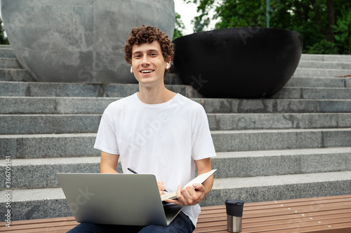 Working outside, freelance concept - European young man in wireless earphones wearing white t-shirt with laptop computer studying outdoor and writing in note book with pencil and listening to music.