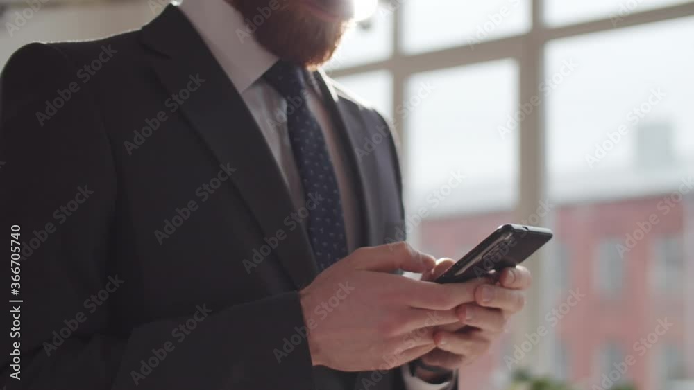 Midsection shot of businessman in formal suit standing in sun-filled office and typing on smartphone while surfing the mobile Internet during workday Midsection shot of businessman in formal suit standing in sun-filled office and typing on smartphone while surfing the mobile Internet during workday