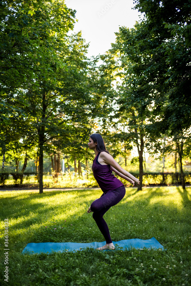 A brunette girl performs the Utkatasana asana in nature at sunset