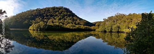 Early morning panoramic view of a calm creek with beautiful reflections of blue sky, mountains and trees on water, Cockle Creek, Bobbin Head, Ku-ring-gai Chase National Park, New South Wales Australia