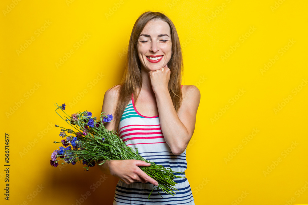 Smiling young girl with closed eyes holding wildflowers on yellow background.