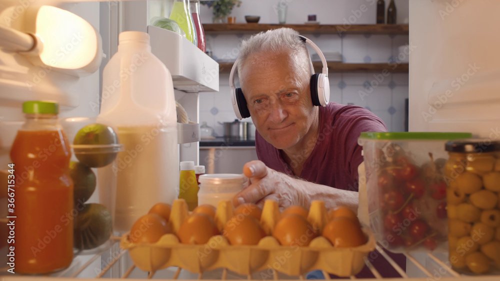 Mature man in headphones taking yogurt for snack from fridge