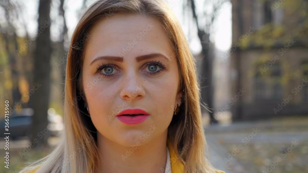 Close up woman in yellow cloak, white blouse stands and looks at the camera in the park, camera moves girl. Girl with pink lipstick on lips outdoors. 4K slow motion footage Close up woman in yellow cloak, white blouse stands and looks at the camera in the park, camera moves girl. Girl with pink lipstick on lips outdoors. 4K slow motion footage