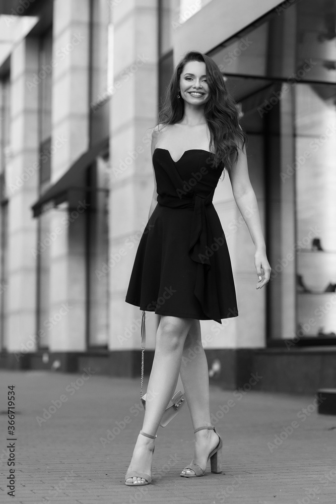 Young beautiful stylish woman in vlack dress, shoes, sunglasses holding handbag standing and posing at city street. Full length outdoor portrait