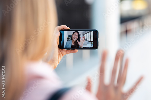 Back view of woman having video conversation with pretty young business woman while staying in the street. photo