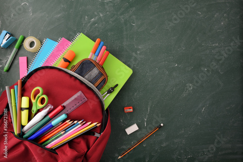 Backpack full of school supplies on green blackboard top view