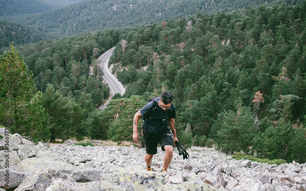 Senderista con bastones en primer plano subiendo por canchal hacia la Bola del Mundo con carretera de montaña al fondo en el Parque Nacional de la Sierra de Guadarrama en Madrid