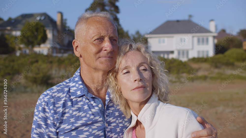 Happy senior couple hugging on sandy beach over country house background