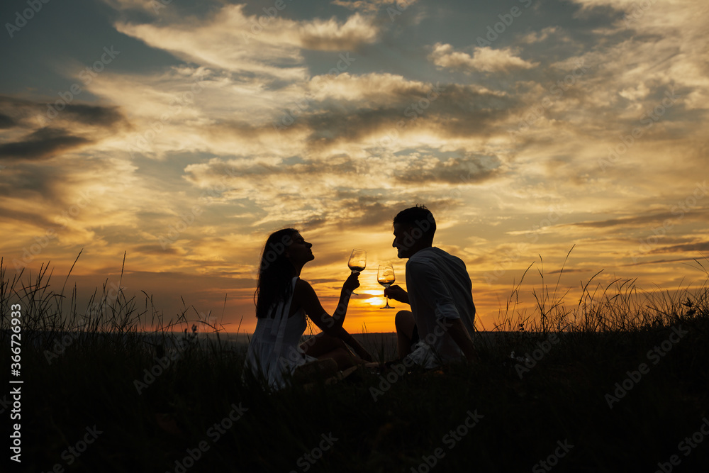 Obraz premium Silhouettes of young man and woman on holidays or honeymoon at amazing sunset sky. Man and woman clanging wine glasses with wine at sunset. Couple in love drinking champagne and celebrating their love