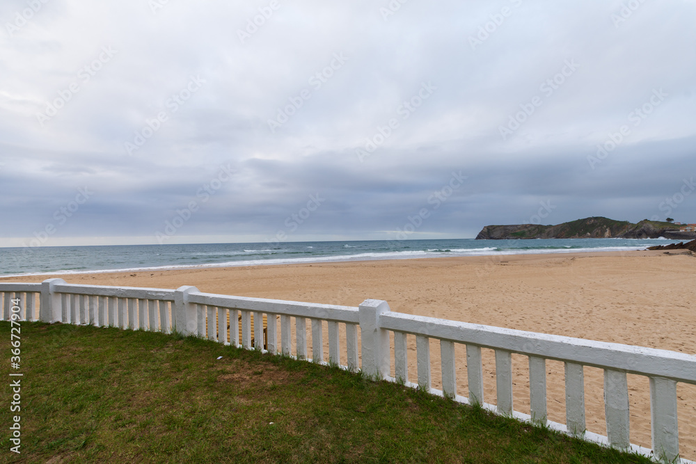 View of white fence on a deserted beach, a cloudy afternoon, with the sea in the background and the green coast, in Comillas, Cantabria, Spain, horizontal