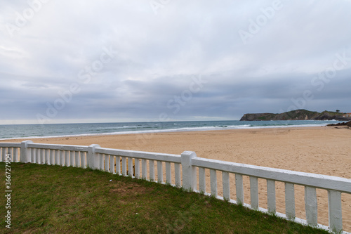 View of white fence on a deserted beach  a cloudy afternoon  with the sea in the background and the green coast  in Comillas  Cantabria  Spain  horizontal