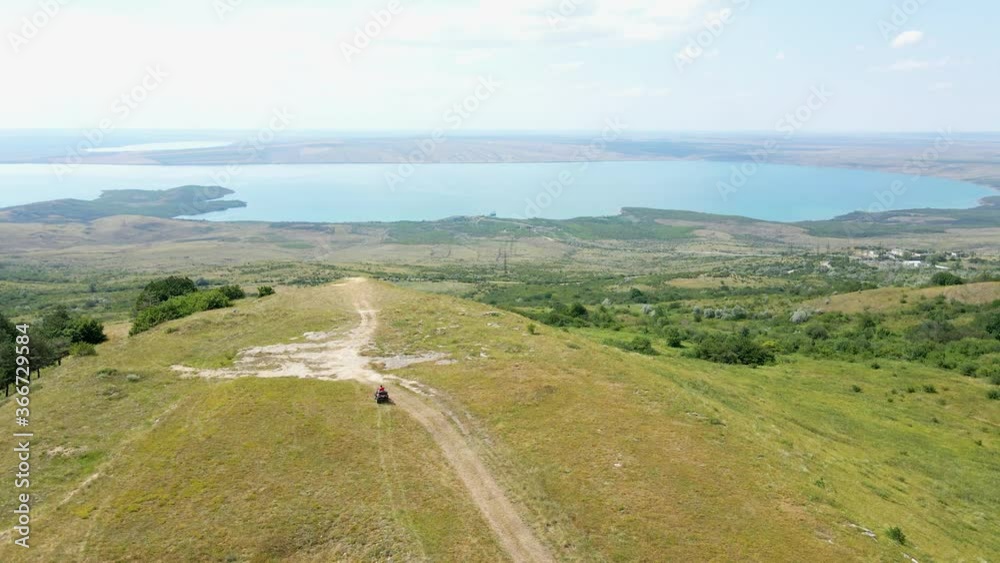 Drone view man in a black cap and red t-shirt on a colored ATV rides on a green mountain hill in summer Drone view man in a black cap and red t-shirt on a colored ATV rides on a green mountain hill in summer
