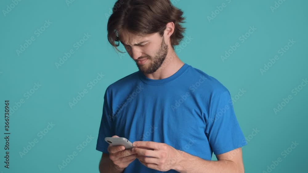 Attractive bearded guy in blue t-shirt disappointedly using smartphone waving no gesture over colorful background