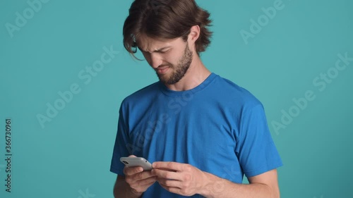 Attractive bearded guy in blue t-shirt disappointedly using smartphone waving no gesture over colorful background