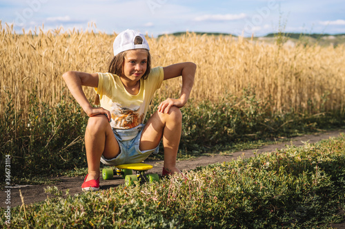 A little girl in a denim jumpsuit sits on a skateboard in the middle of a wheat field, a cheeky naughty teen