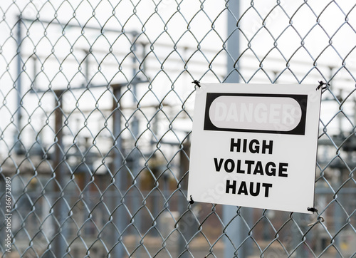 A bilingual faded High Voltage sign on a chain link fence, on a dark cloudy day. It is slightly cooked. The red on the sign is faded and almost invisible. A substation is in the background.