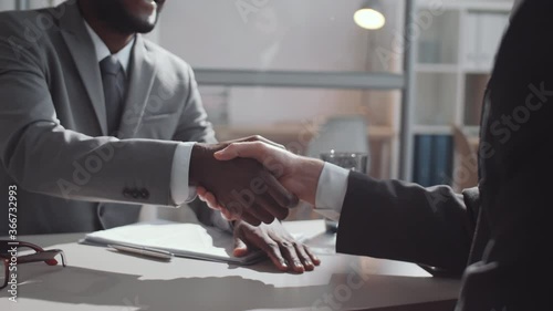 Midsection arc shot of African American and Caucasian male colleagues in formalwear shaking hands and discussing business while sitting at desk in office