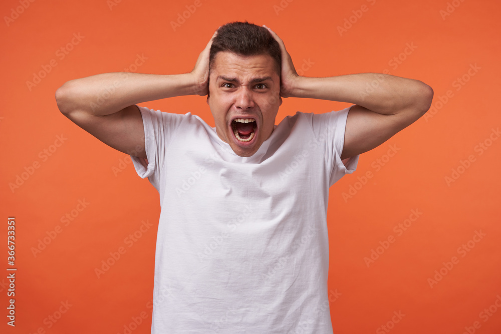 Irritated young brown-eyed short haired male covering his ears with raised palms and keeping mouth opened while screaming excitedly, isolated over orange background Irritated young brown-eyed short haired male covering his ears with raised palms and keeping mouth opened while screaming excitedly, isolated over orange background