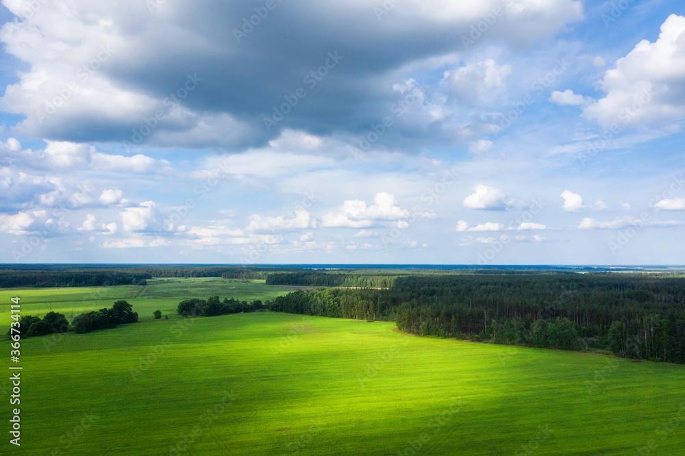 Sunny aerial view of forest and meadow. Summer landscape. A lot of white clouds on sky. Sunny beams  throught clouds. Various shades of green color.