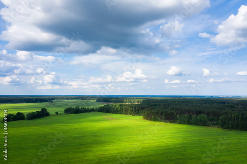 Sunny aerial view of forest and meadow. Summer landscape. A lot of white clouds on sky. Sunny beams throught clouds. Various shades of green color.