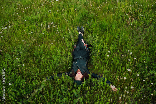 A young woman of 25-30 years old with curly hair, in a dress, a black kodan jacket and a diadem with feathers lies on the grass. photo