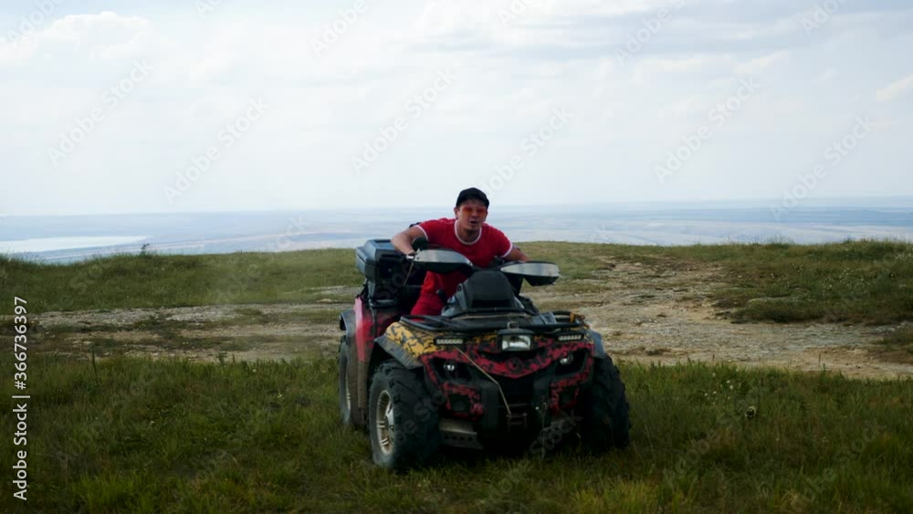 man in a black cap and red t-shirt on a colored ATV rides on a green mountain hill in summer
