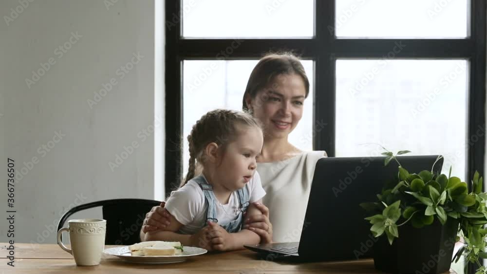 Young american mother and cute daughter have video call at table with laptop in apartment room iroi. Beautiful woman and little girl talking with smiles and looking at computer screen while sitting at Young american mother and cute daughter have video call at table with laptop in apartment room iroi. Beautiful woman and little girl talking with smiles and looking at computer screen while sitting at