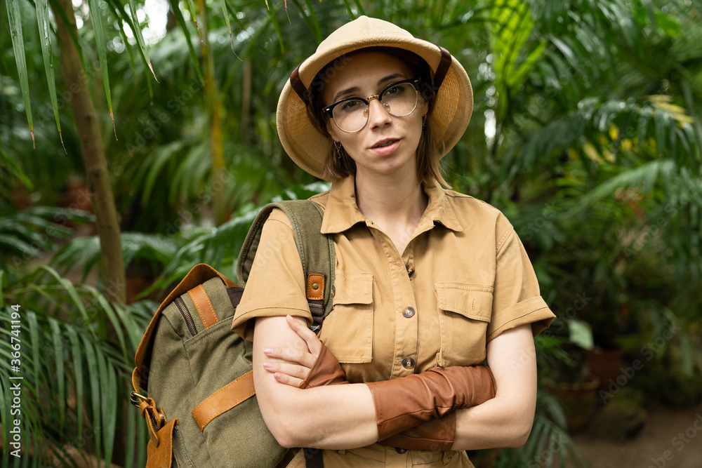Close up portrait of woman botanist dressed in safari style in greenhouse. Naturalist in khaki clothes, leaver gloves with backpack looking at camera, arms crossed, surrounded by palms.  
