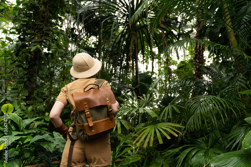Woman botanist dressed in safari style in greenhouse, back view. Naturalist in khaki clothes, leaver gloves with backpack walks in the rainforest surrounded by palms. Jungle tourist photo