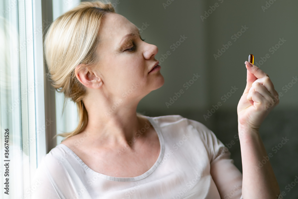 Obraz premium Women and drug capsule. Portrait close. The girl's face, blond hair, beige dress. Capsule in the mouth, capsule in the hand. Dietary supplement. Women 40 years old looks so young. 
