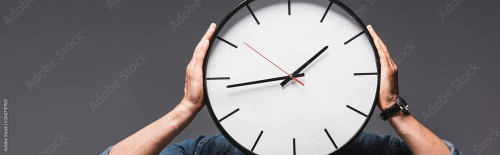 Panoramic crop of man holding clock near face isolated on grey, concept of time management Panoramic crop of man holding clock near face isolated on grey, concept of time management