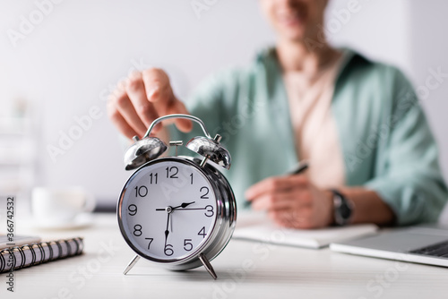 Selective focus of man pulling hand to alarm clock near laptop and notebooks on table, concept of time management photo