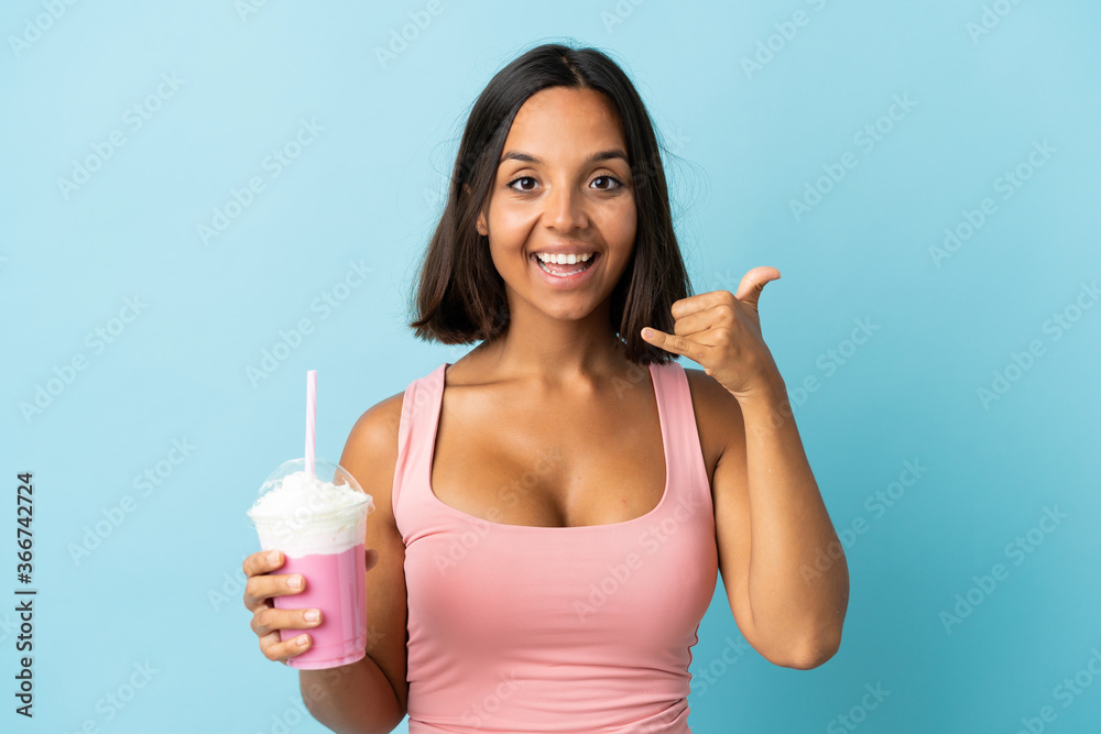 Young woman with strawberry milkshake isolated on blue background making phone gesture. Call me back sign Young woman with strawberry milkshake isolated on blue background making phone gesture. Call me back sign