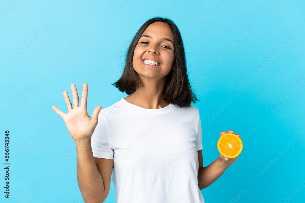 Young asian girl holding an orange isolated on blue background counting five with fingers Young asian girl holding an orange isolated on blue background counting five with fingers