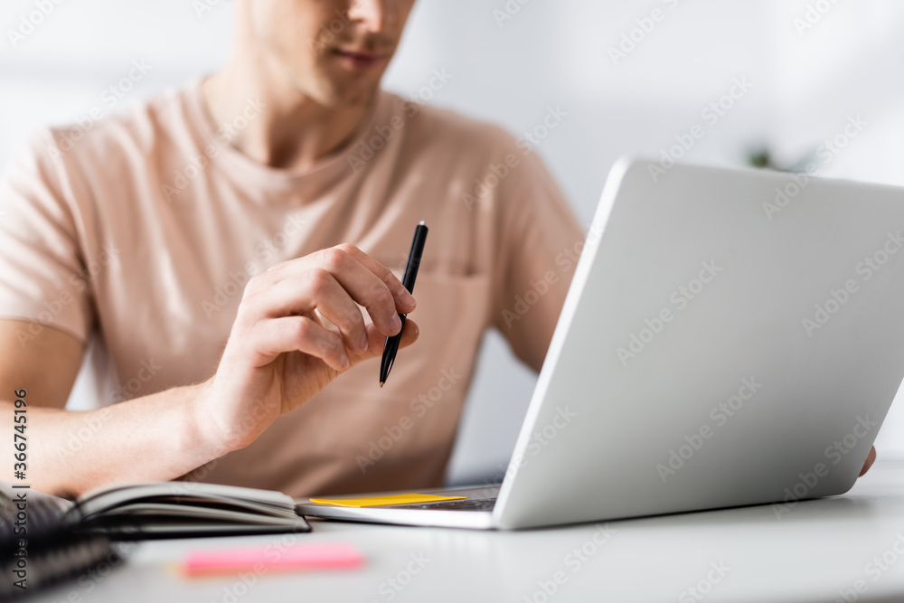 Cropped view of freelancer holding pen and using laptop on table, earning online concept Cropped view of freelancer holding pen and using laptop on table, earning online concept