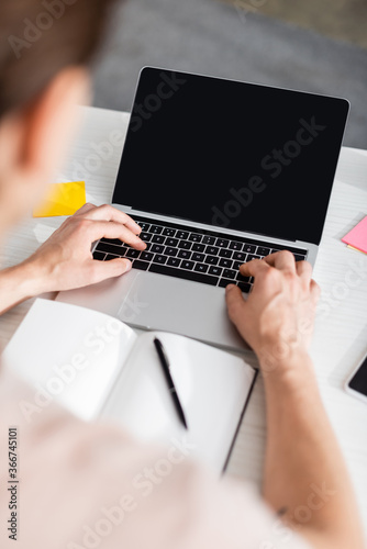 Cropped view of man typing on laptop with blank screen, sitting at table at home, earning online concept