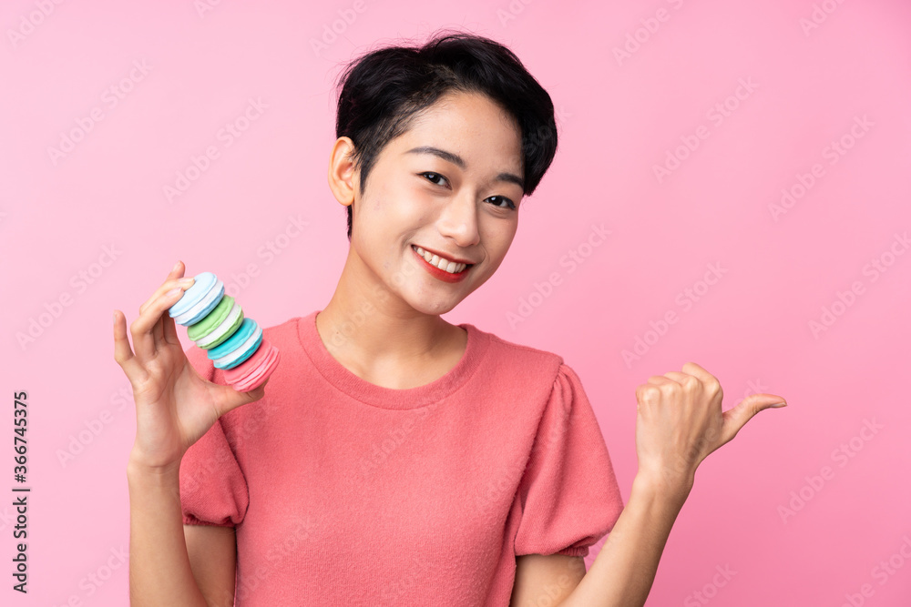 Young Asian girl over isolated pink background holding colorful French macarons and pointing side Young Asian girl over isolated pink background holding colorful French macarons and pointing side