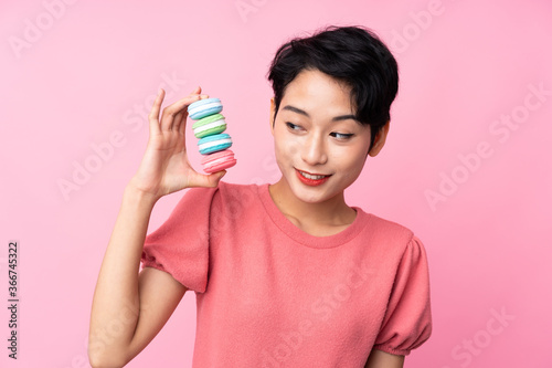 Young Asian girl over isolated pink background holding colorful French macarons and with happy expression