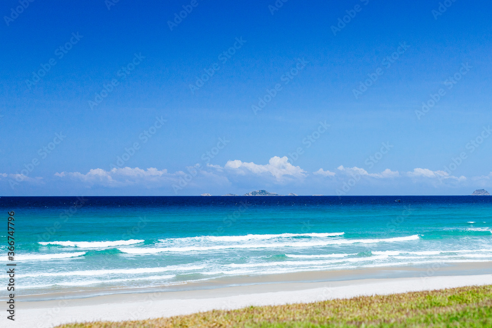 Landscape view of the sandy sea beach. Calm waves, foam. Blue sky with white clouds. Summer vacation, travel. Background, copy space.
