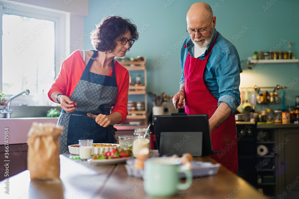 A cheerful couple in their fifties in their kitchen are preparing a strawberry tart. They are looking for the recipe on a digital tablet