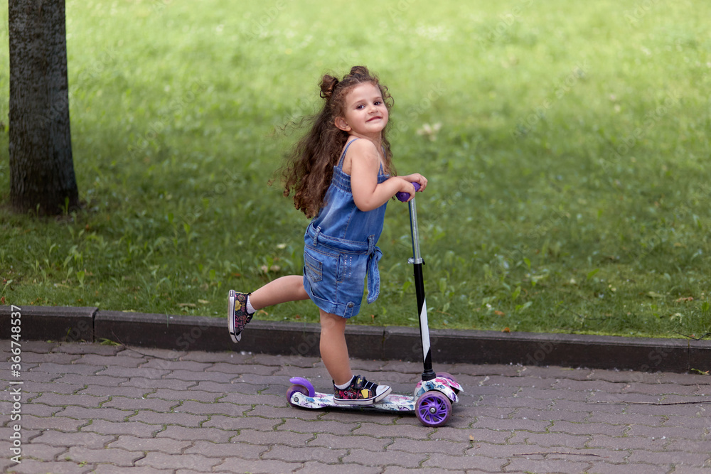 Naklejka premium Beautiful little girl rides a scooter in the park. Beautiful brunette girl.