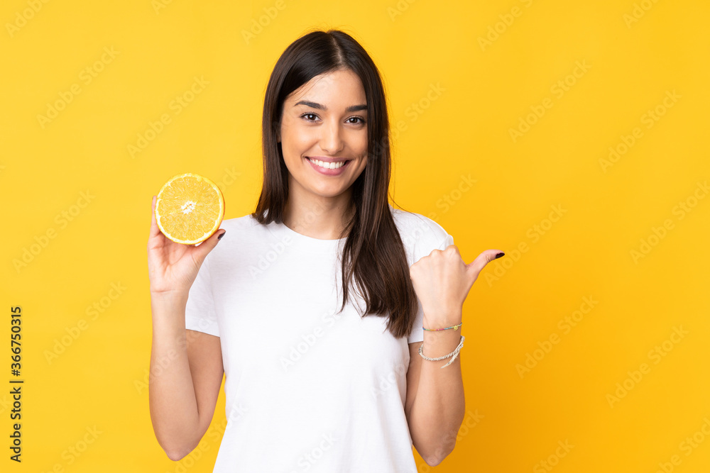 Young caucasian woman holding an orange isolated on yellow background pointing to the side to present a product Young caucasian woman holding an orange isolated on yellow background pointing to the side to present a product