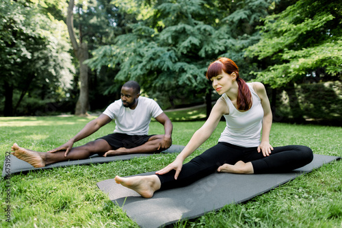 Multiethnic couple doing yoga in nature. Young Caucasian woman performs yoga asanas outdoors in a park, together with her handsome African boyfriend behind. Yoga, fitness, sports, healthy lifestyle © sofiko14