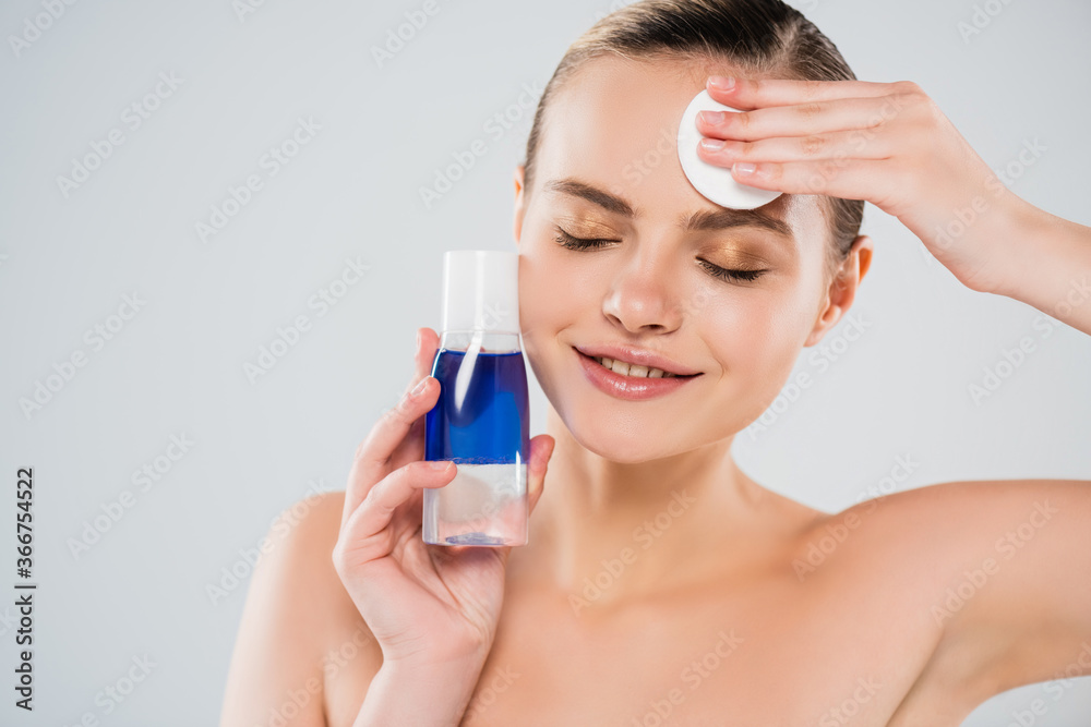 happy and naked woman holding bottle while applying toner with cotton pad isolated on grey happy and naked woman holding bottle while applying toner with cotton pad isolated on grey