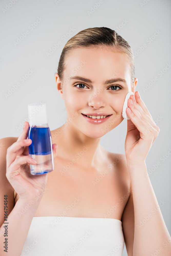 happy woman holding bottle while applying toner with cotton pad isolated on grey happy woman holding bottle while applying toner with cotton pad isolated on grey