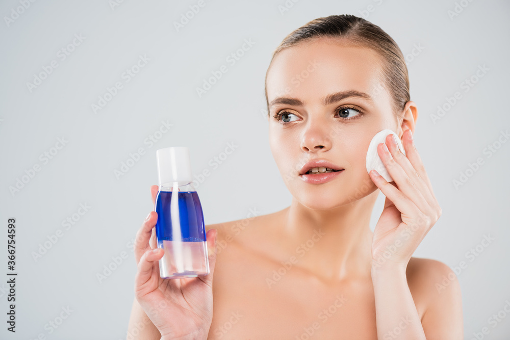 naked woman holding bottle while applying toner with cotton pad isolated on grey naked woman holding bottle while applying toner with cotton pad isolated on grey