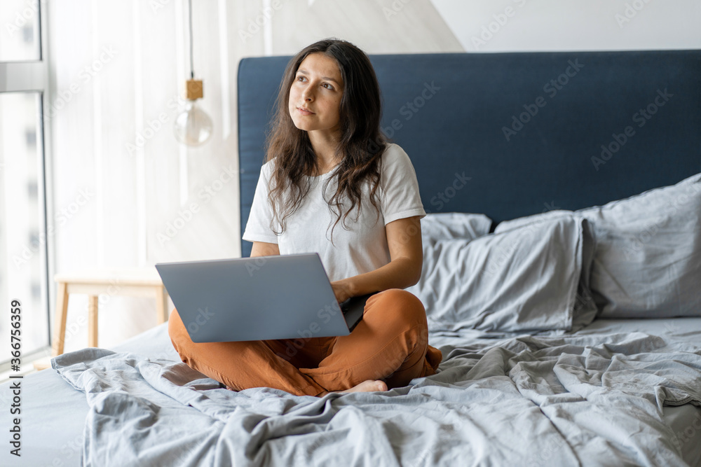 Beautiful young brunette girl with a laptop sitting on the bed. Stylish modern interior. A cozy workplace. Shopping on the Internet Beautiful young brunette girl with a laptop sitting on the bed. Stylish modern interior. A cozy workplace. Shopping on the Internet